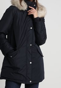 Schwarze gepolsterte Winterjacke mit einem Kragen aus Kunstfell, Druckknöpfen, Fronttaschen und gerippten Bündchen. Glatte Textur mit taillierter Silhouette.