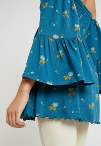 Sininen kukallinen toppi, jossa on kellohihat, valkoisia ja keltaisia kuvioita, teksturoitua kangasta ja aaltoileva helma.