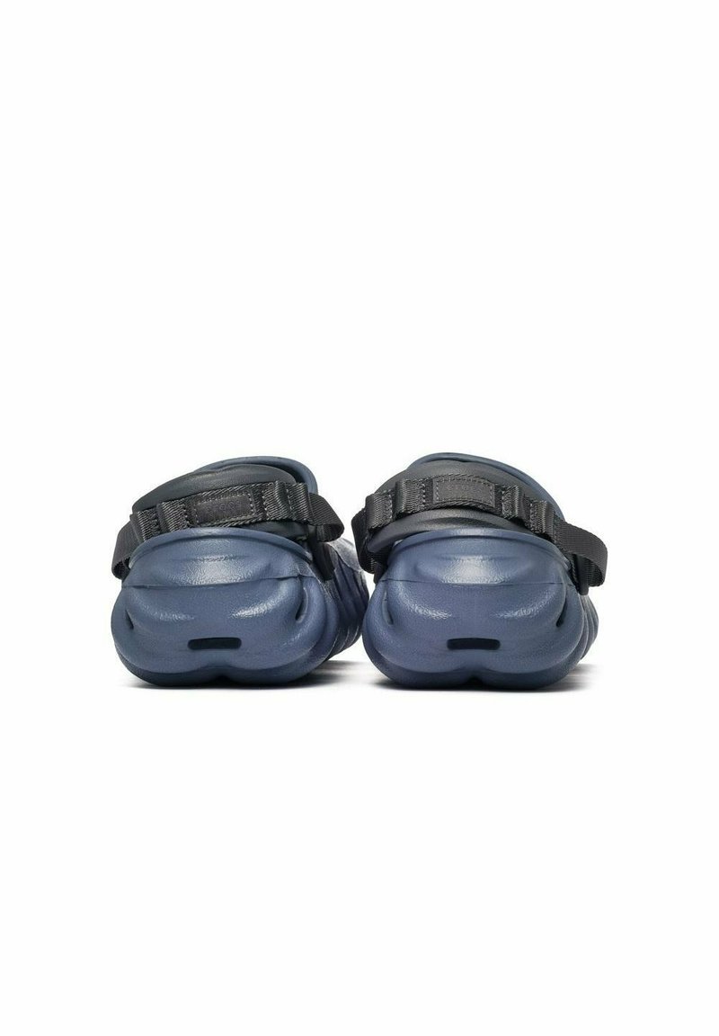 Vista trasera de sandalias azul marino tipo slip-on con suelas gruesas y correas ajustables negras sobre un fondo blanco.