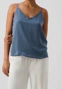 Top blu senza maniche con scollatura ondulata, realizzato in tessuto leggero con una consistenza liscia. Indossato con pantaloni bianchi larghi.