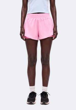 Person iført lyserøde atletikshorts, hvide sokker, sorte løbesko og en hvid cropped top, stående mod en ensfarvet baggrund.