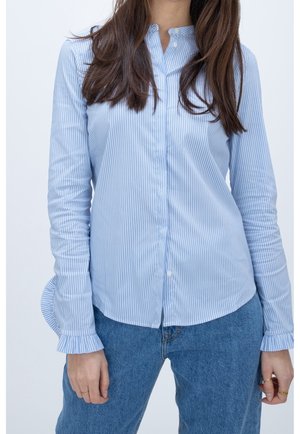 Kobieta ubrana w jasnoniebieską i białą koszulę w paski z falbankami na mankietach oraz niebieskie dżinsy, stoi na jednolitym tle.