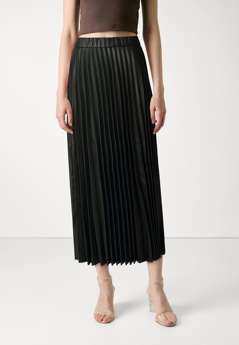 Femme portant une jupe midi plissée noire taille haute associée à des sandales à talons hauts à brides transparentes sur un fond uni.