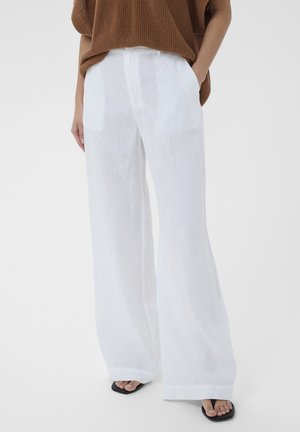 Person wearing white wide-leg pants, brown knit sweater, and black open-toe sandals with toes visible, standing with one hand in pocket.