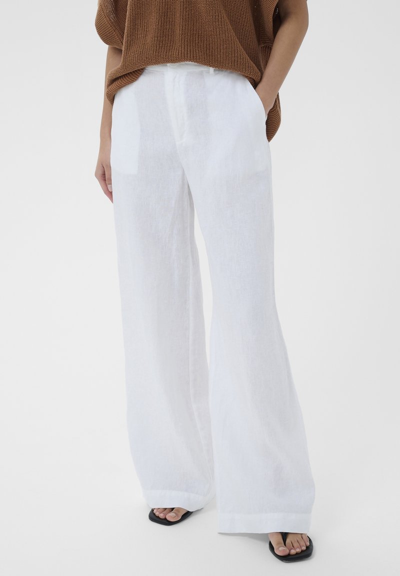 Personne portant un pantalon blanc à jambes larges, un pull en tricot marron et des sandales noires ouvertes laissant apparaître les orteils, debout avec une main dans la poche.