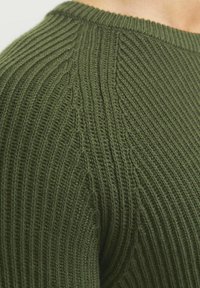 Detailní záběr na zelený svetr z ribového úpletu, ukazující propracovaný vertikální a diagonální šev poblíž ramene a kolem výstřihu.