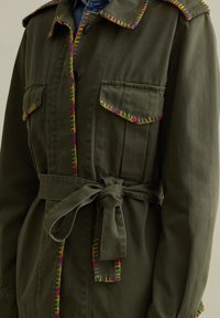 Giacca verde oliva con cuciture multicolori lungo il colletto e le tasche, caratterizzata da una vita con cintura e due tasche sul petto.