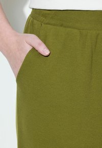 Olijfgroene textuur broek met een zijzak, voorzien van een geribbeld patroon en een elastische tailleband, getoond met een hand die in de zak rust.