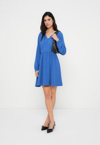 Μπλε φόρεμα με μακριά μανίκια, σφηκοφωλιά στη μέση και φουσκωτή φούστα, σε συνδυασμό με μαύρη τσάντα πουά και μαύρες μυτερές γόβες.