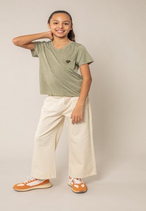 Fille souriante aux cheveux bruns attachés en queue de cheval, portant un t-shirt vert, un pantalon large crème et des baskets orange, posant avec une main derrière la nuque.