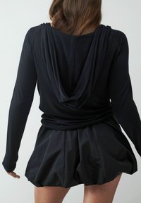 Schwarzes Langarmoberteil mit einer drapierten Kapuze und lockerer Passform, kombiniert mit einem schwarzen strukturierten Rock in blousonform. Einfaches, monochromatisches Design.