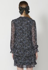 Rochie florală neagră cu mâneci lungi din material transparent, straturi cu volane și detaliu cu fundă la spate. Tipare florale albastre și albe pe întreaga țesătură.