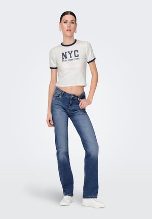 T-shirt blanc court avec des bordures bleu marine et le texte "NYC", associé à un jean bleu évasé. Le mannequin se tient contre un fond uni, portant des baskets blanches.