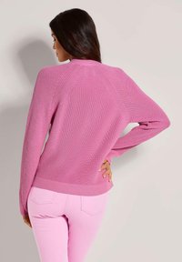 Vrouw met lang donker haar die een roze gebreide trui en lichtroze broek draagt, met één hand op haar heup, met de rug naar de kijker gekeerd.