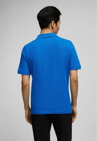 Polo bleu en tissu texturé, à manches courtes et avec col. La chemise présente un ourlet droit et est associée à un pantalon noir.