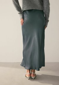 Satin grüner Maxi-Rock mit glatter Textur und fließendem Schnitt, kombiniert mit silbernen Absatzsandalen und einem grauen Strickpullover.