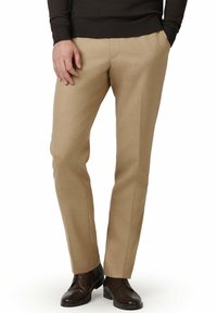 Beżowe, dopasowane spodnie wykonane z gładkiej tkaniny, o wąskim kroju, z przednimi kieszeniami i czystą linią designu. Noszone z ciemnobrązowymi butami.
