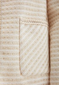 Kremowa tkanina z teksturą, z złotymi nitkami, z plecionym wzorem. Widoczna jest mała kieszonka, wykończona plecionym brzegiem.