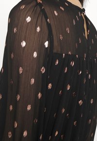 Vestido negro con tela semitransparente que presenta lunares reflectantes en oro rosa. Un cierre de botón en la parte posterior añade un detalle sutil. Diseño fluido.