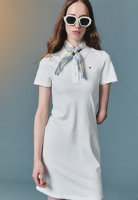 Balta polo suknelė su apykakle ir mygtukų eilių. Papuošta mėlyna ir kreminės spalvos šaliku, kuris surištas aplink kaklą, ir didelėmis baltomis saulės akimis.