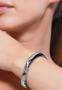 Bracciale rigido in metallo argentato con un design liscio e curvo, che mostra una finitura lucida e riflessi sottili sulla superficie.