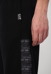Crne atletske hlače s teksturiranim dijelom na kojem se ponavlja tekst "EA7 Emporio Armani". Materijal je mekan i rastezljiv. Vidljiv je oznaka s logotipom.