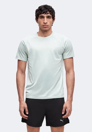 Junger Mann mit lockigem Haar, der ein hellgraues Kurzarmhemd und schwarze Sportshorts trägt, steht vor einem schlichten Hintergrund.