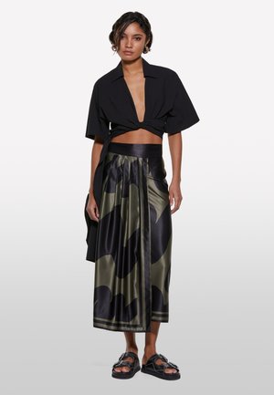 Chemise noire raccourcie avec un décolleté en V profond, associée à une longue jupe satin olive foncé présentant un motif noir audacieux, portée avec des sandales noires.