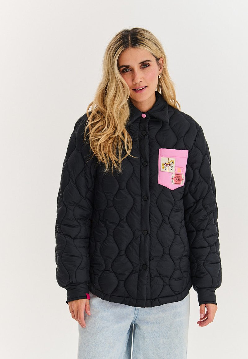 Žena s dlouhými blond vlasy má na sobě černou prošívanou bundu s růžovou náprsní kapsou zdobenou nášivkou koně a designem lístku, kterou kombinuje se světlými džínami.