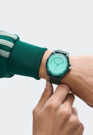 Zaļš pulkstenis ar apaļu piparmētru krāsas seju, stundu rādītājiem un melnu gumijas siksniņu. Rokas pielāgo pulksteni pie plaukstas, uz kuras vilkta zaļa garajā piedurknē tops.