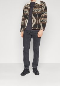 Gekraagde jas met gemengde geometrische patronen in beige, bruin en zwart, gecombineerd met een zwart t-shirt en donkere jeans; zwarte schoenen.