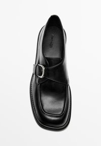 Zwarte leren schoen met een vierkante neus, kruisband en metalen gespsiering. Soepele textuur en gedetailleerde stiksels langs de zool.