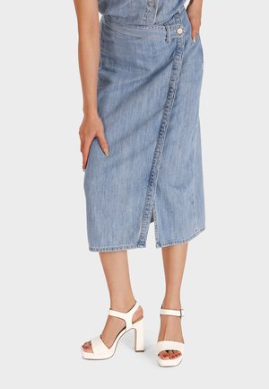 Gonna midi in denim di colore azzurro chiaro con design avvolgente, caratterizzata da uno spacco frontale e chiusura con bottoni. Abbinata a sandali bianchi con tacco a blocco.