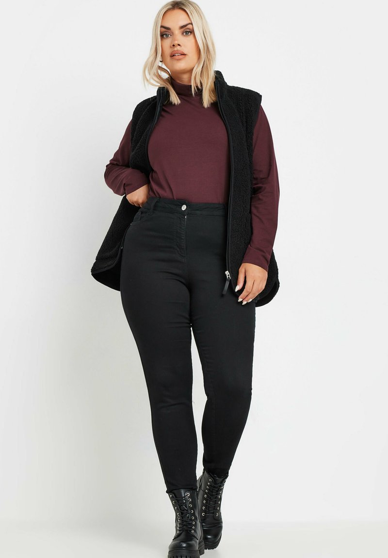 Czarna polarowa kamizelka nałożona na bordowy długi rękaw, w połączeniu z czarnymi obcisłymi dżinsami i czarnymi lace-up botkami za kostkę.