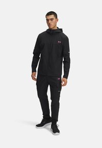 Zwarte sportjack en broek van lichtgewicht stof met roze accenten, ritssluiting aan de voorkant, zakken en een capuchon. Heeft een strakke uitstraling.