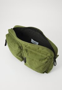 Green canvas bag with two front pockets, zip closure, and black interior lining. Features stitched detail and branded label inside.