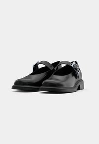Black synthetic shoes with a glossy finish, featuring a wide strap and metallic buckle detail, low block heel, and rounded toe design.