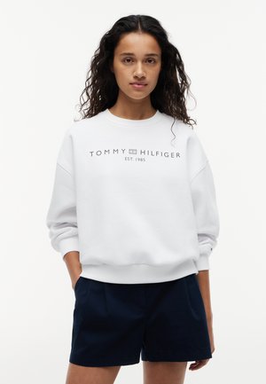 Ung kvinne med krøllete hår som har på seg en hvit Tommy Hilfiger-genser og marineblå shorts, stående med den ene hånden i lommen på hvit bakgrunn.