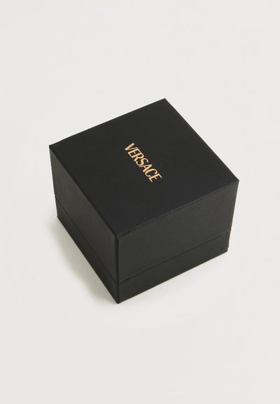 Μαύρο κουτί κοσμημάτων με χρυσό λογότυπο "VERSACE" στο καπάκι, τοποθετημένο σε απλό, ανοιχτό φόντο.