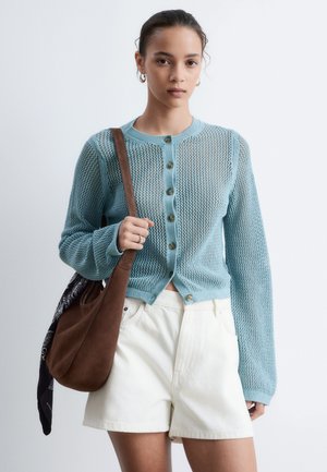 Junge Frau mit hellblauer Strickjacke, weißen High-Waist-Shorts und einer braunen Umhängetasche mit schwarzem Schal.
