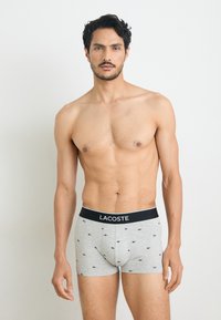 Gray boxer shorts with black Lacoste logo waistband, featuring small blue alligator patterns. Soft fabric, fitted design, mid-thigh length.
