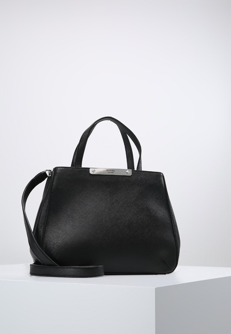 Sac à main Guess en cuir noir avec poignées courtes et bandoulière détachable sur un piédestal blanc.