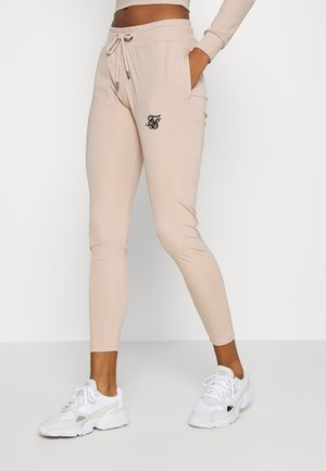 Jogging beige slim-fit avec taille à cordon et poches latérales, porté avec des baskets blanches et un top cropped à manches longues, debout sur un fond blanc.