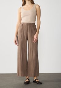 Lichtbeige tanktop gecombineerd met geplooide, wijd uitlopende bruine broek. De broek heeft een elastische tailleband en zachte, gestructureerde stof. Zwarte platte schoenen maken de look compleet.