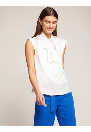 Mujer con blusa blanca sin mangas con lazo y pantalones azul brillante, sonriendo con los ojos cerrados sobre un fondo liso.