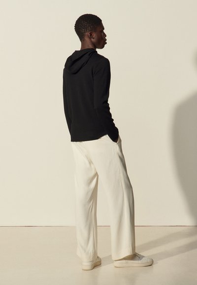 Sweat à capuche noir, pantalon large crème et baskets claires. Tissu lisse, coupe décontractée et design minimal. Fond neutre.