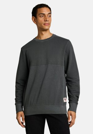Mann trägt ein dunkelgraues Sweatshirt mit Rundhalsausschnitt und schwarze Hose, steht vor einem schlichten hellen Hintergrund.