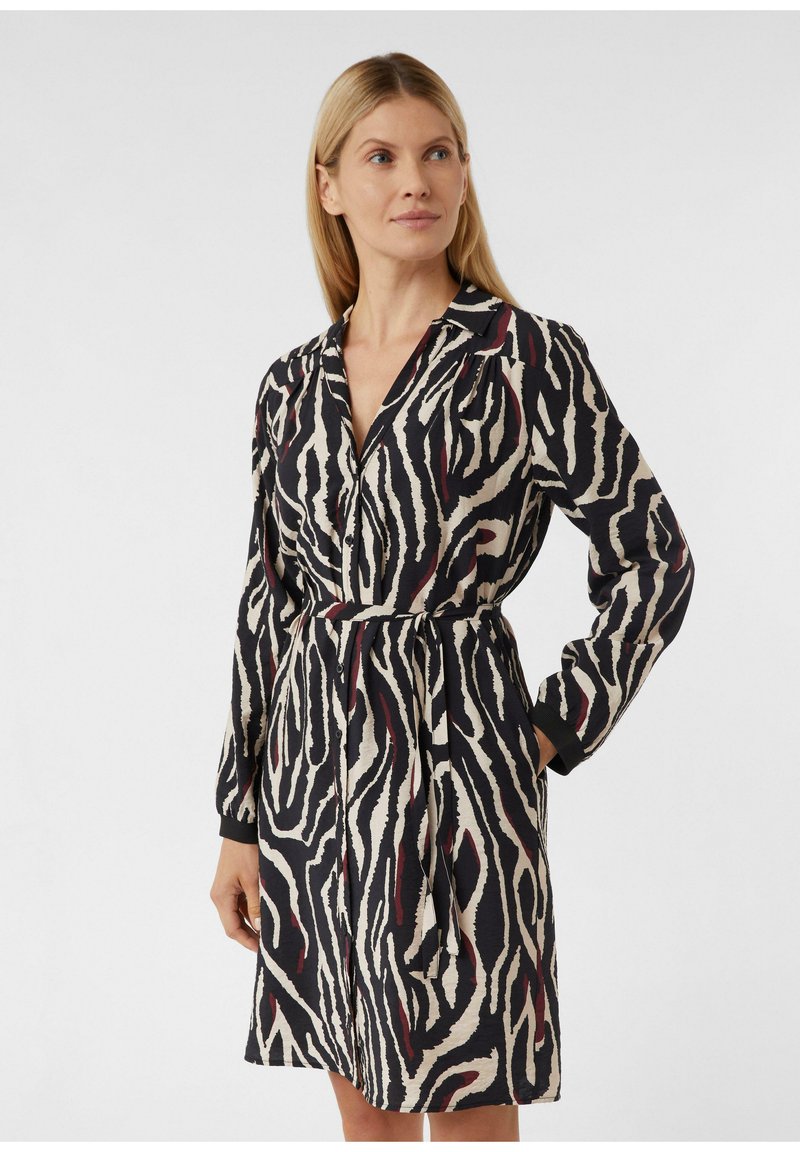 Vrouw in een jurk met lange mouwen en knopen, bedekt met zwart, wit en donkervroeg zebrapatroon, met een bijpassende tailleband, staande met één hand in de zak.