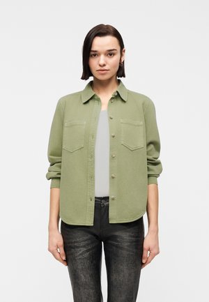 Young person with short dark hair wearing an olive green buttoned shirt over a light gray top and black textured pants, standing against white background.