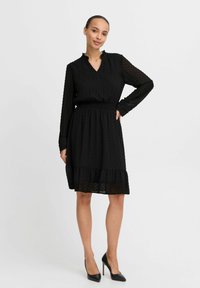 Robe noire avec un motif à pois, manches longues, décolleté en V, col volants et ourlet à étages. Taille plissée pour un ajustement structuré.